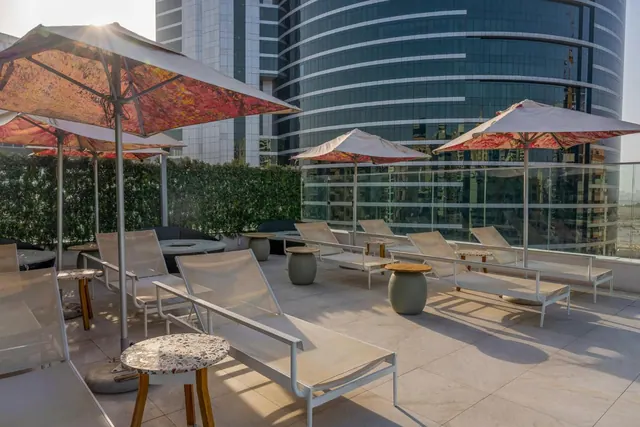 A terrace with white sun loungers and shade umbrellas sits beside a glass railing, with tall glass towers rising behind it.