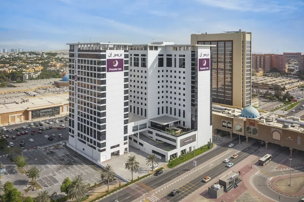 Aerial view of a white Premier Inn hotel tower with purple signage beside a busy road lined with palm trees.