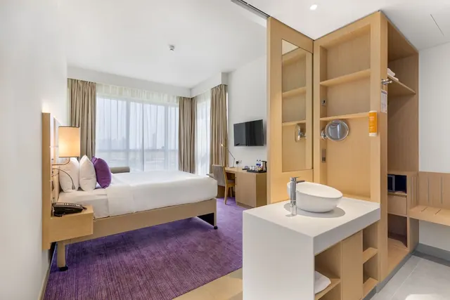 A hotel bedroom with a double bed with purple cushions, a wall-mounted television and desk, a large window with sheer curtains, and a wood-panelled wardrobe with a white vessel sink in the foreground.