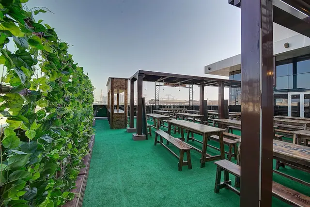 An outdoor rooftop seating area with wooden tables and benches under a pergola, a green artificial turf floor, and a climbing plant wall on the left.