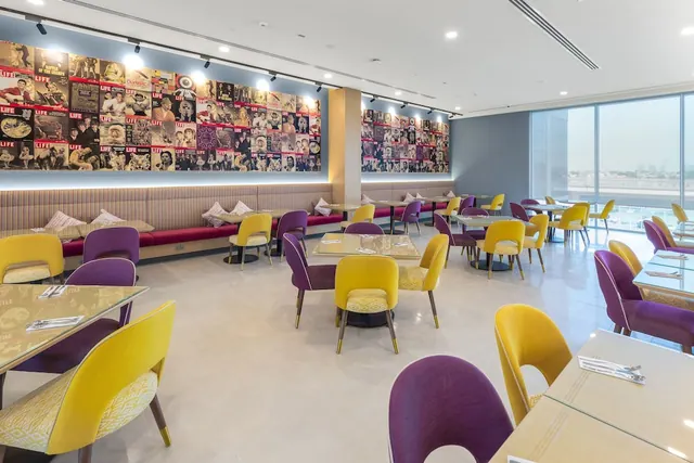 A restaurant dining room with yellow and purple chairs, banquette seating along a wall covered in vintage Life magazine covers, and large windows looking out onto distant buildings.