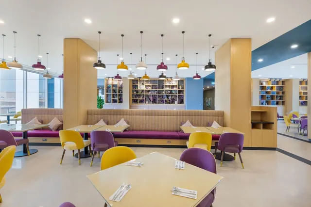 A dining room with booth seating with purple cushions, yellow and purple chairs, pendant lamps hanging above the tables, and wooden shelving.