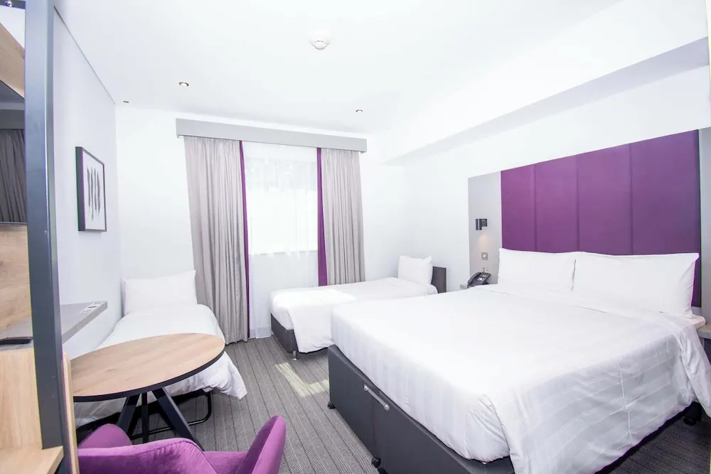 A hotel bedroom with a double bed, a single bed, a round table with a purple chair, and a window with sheer and gray curtains.