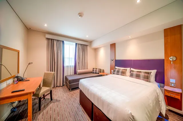 A hotel bedroom with a made double bed under a purple headboard, a bench seat by the curtained window, and a wooden desk with a chair.