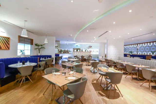 A restaurant dining room with wooden tables, grey chairs, blue banquette seating and a curved white ceiling, set with no diners.
