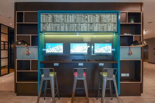 A row of three stools faces a counter with three monitors and keyboards, set against a backlit bookshelf and a teal-framed wall niche.