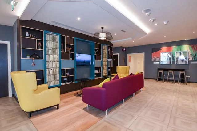 A lounge with yellow armchairs, a magenta sofa, shelving with a wall-mounted television, and a bar counter with stools in the background.