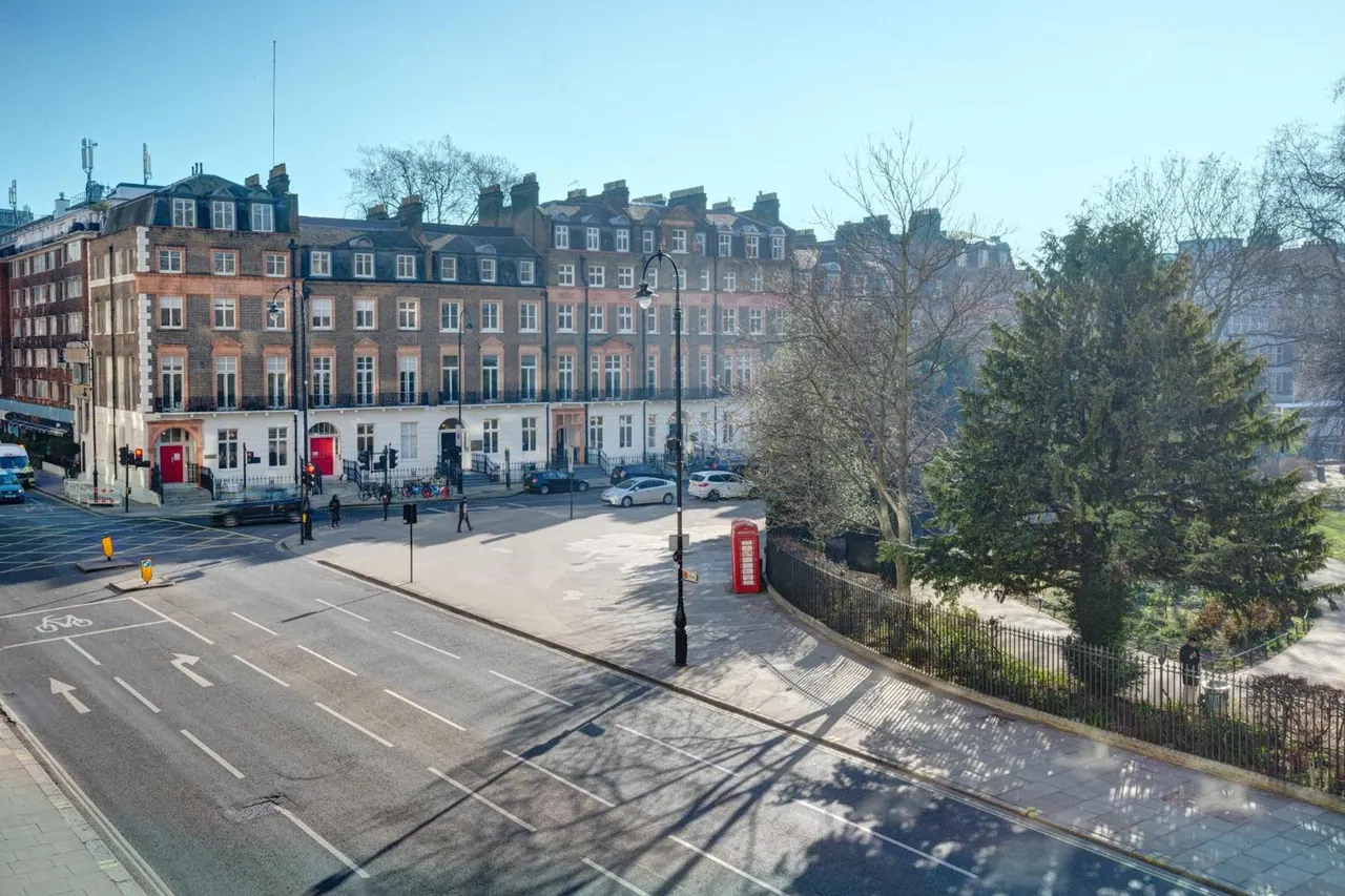 Elevated view over a road past a row of brick terraced houses, a red telephone box and a tree-lined garden railing.