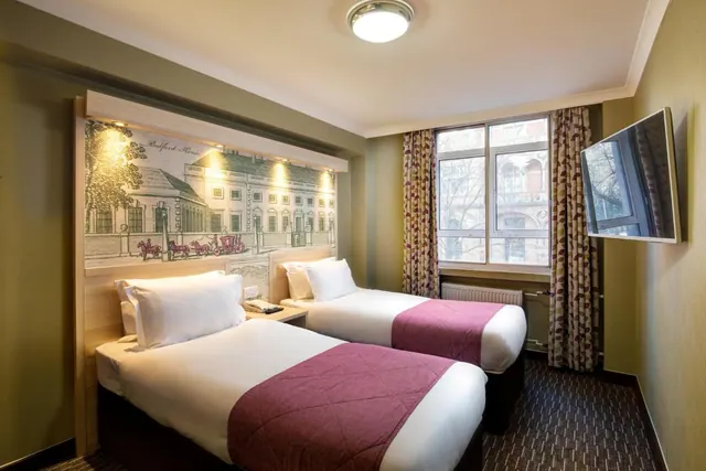 A twin bedroom with two single beds, a painted mural headboard, a wall-mounted television and a window looking onto a building across the street.