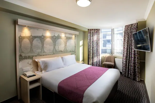 A double bed with white bedding and a magenta throw sits in front of a bridge mural headboard, with a bay window, armchair and wall-mounted television.