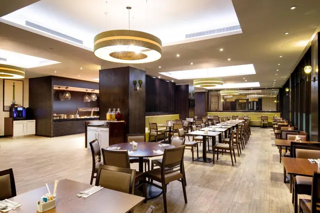 A restaurant dining room with rows of wooden tables and chairs, large pendant lights, green banquette seating, and a buffet counter with a coffee machine on the left.