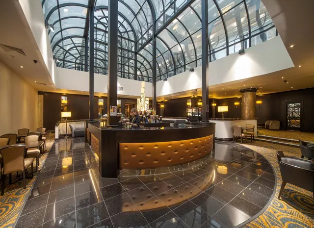 A curved bar with a tufted leather front stands beneath a glass domed ceiling in a hotel lounge, with a bartender behind the counter.
