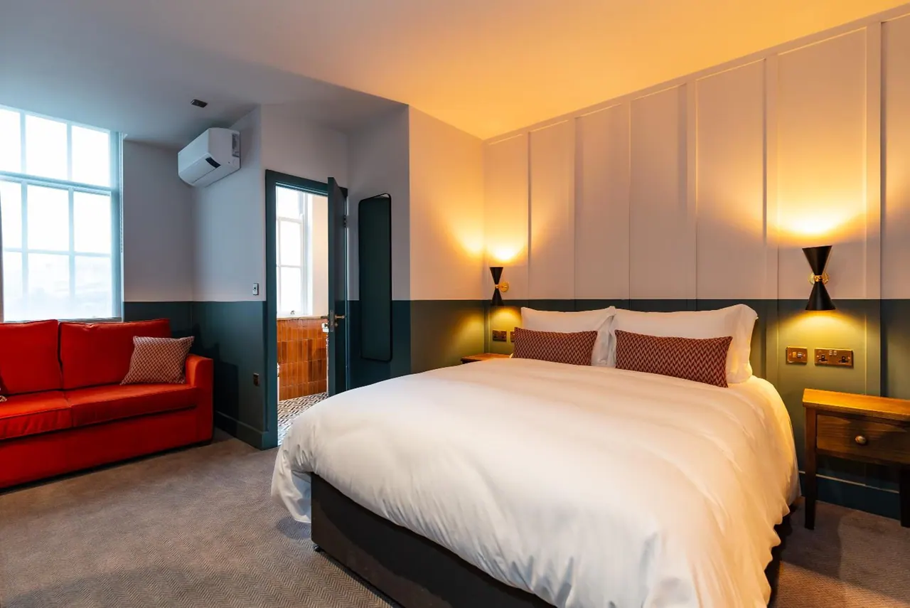 A bedroom with a made double bed, a red sofa by the window, a wall-mounted air conditioning unit, and an open door to a tiled bathroom.