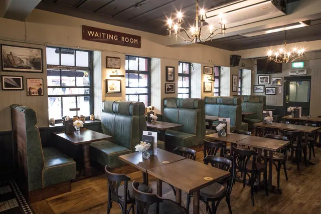A dining room with green leather booth seating, wooden tables and bentwood chairs under chandeliers, with a Waiting Room sign on the wall.