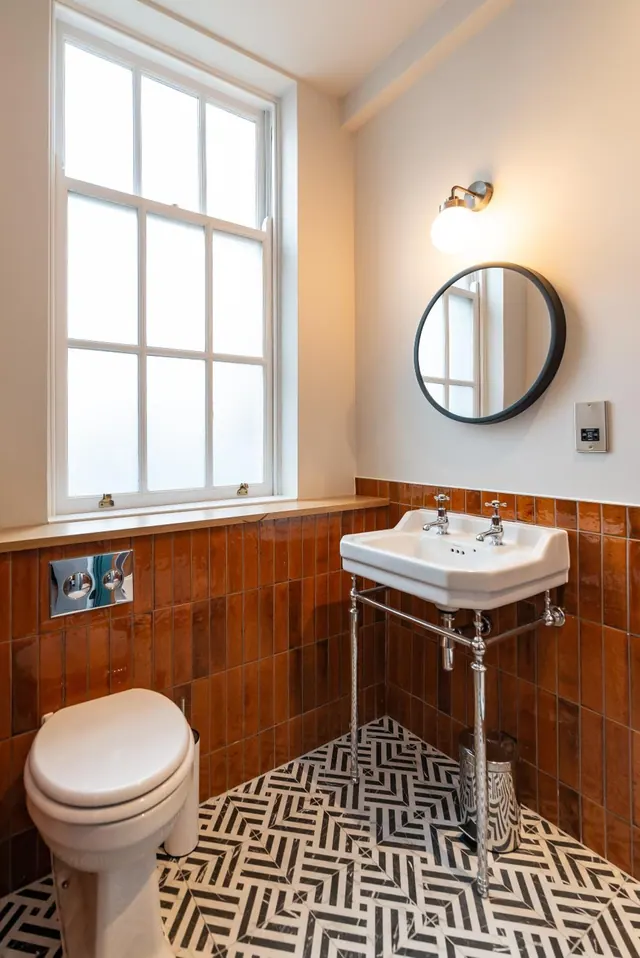 A bathroom with a frosted sash window, glossy orange wall tiles, a pedestal basin beneath a round wall mirror, and a toilet on a black and white patterned tiled floor.
