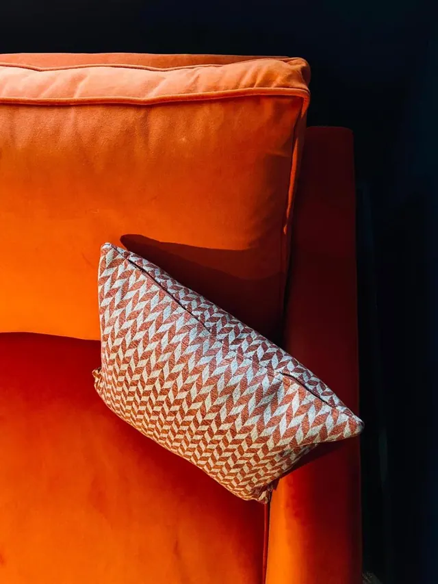 An orange velvet sofa with a patterned cushion is shown against a dark background.