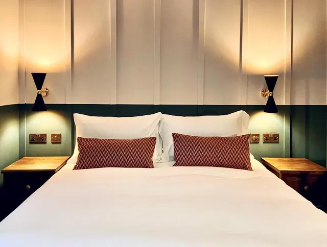 A double bed with white bedding and two red patterned cushions sits between wooden bedside tables under wall lights.