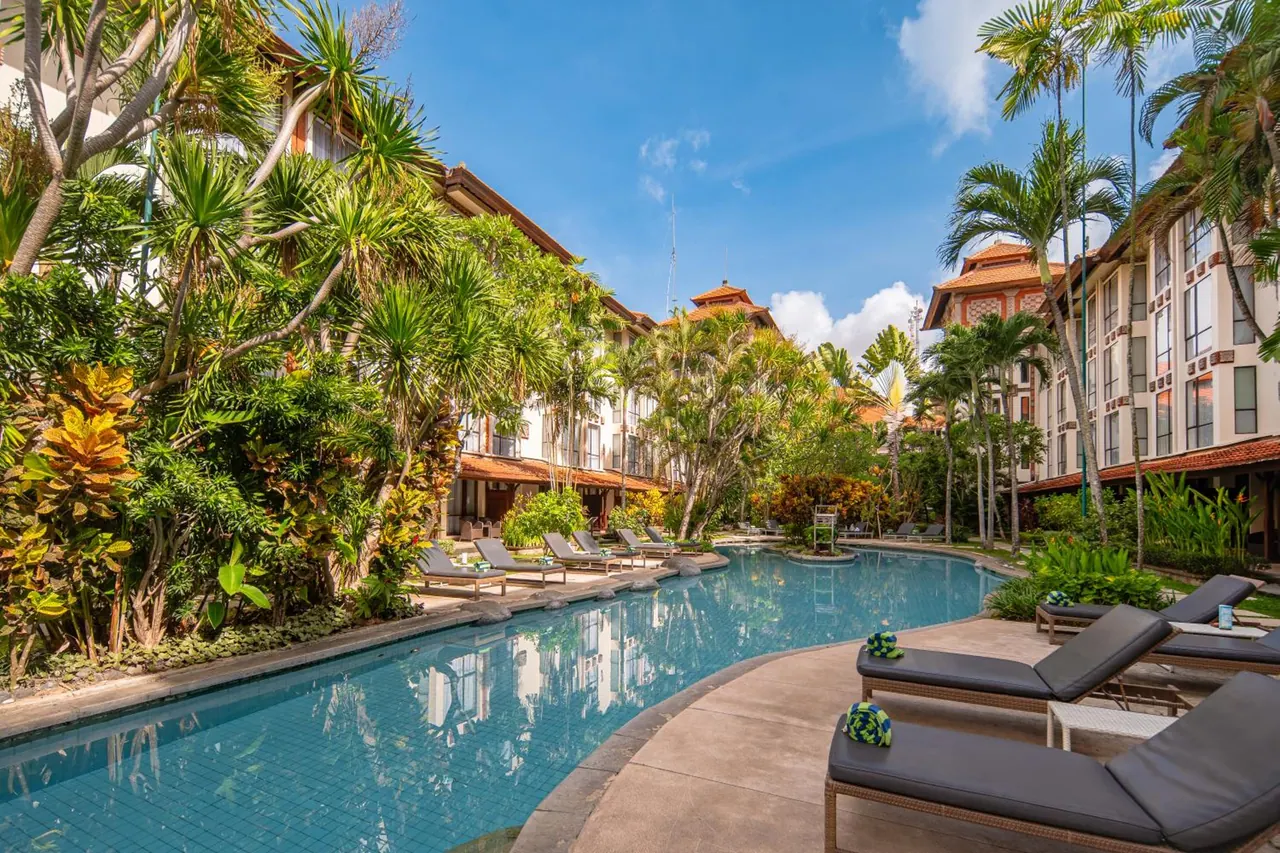 A long curved swimming pool lined with grey sun loungers, surrounded by tropical plants and hotel buildings with red-tiled roofs.