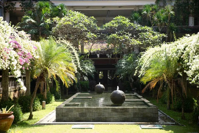 A courtyard garden with a stepped water pool holding two spherical fountains, flanked by palm trees and white flowering shrubs, with a multi-storey building behind.