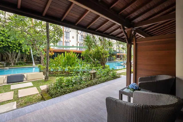 A covered terrace with wicker chairs and a side table overlooks a garden, a swimming pool and a hotel building.