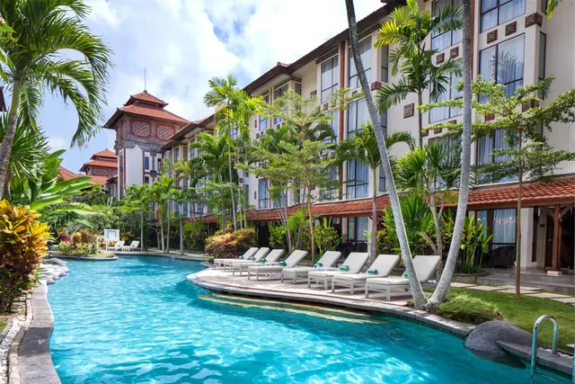 A curved swimming pool with white sun loungers along its edge, with a multi-storey hotel building and palm trees behind it.
