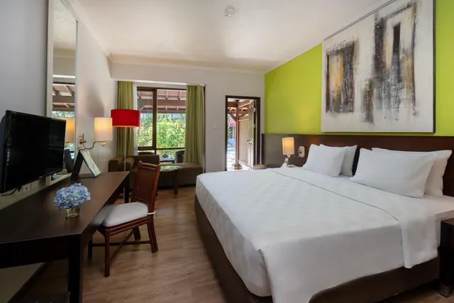 A hotel bedroom with a king bed, a green accent wall, a desk with a television, and a sofa by a window looking onto trees.