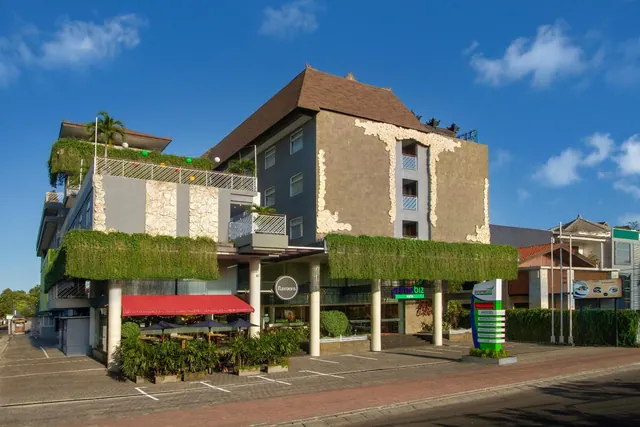 Exterior of a multi-storey hotel with a Balinese-style roof, green planted walls, a red awning over an outdoor dining area with umbrellas, and an empty parking lot in front.