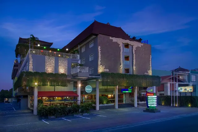 Exterior of a multi-storey hotel at dusk with illuminated signage, a covered restaurant terrace with outdoor tables, and an empty parking area in front.