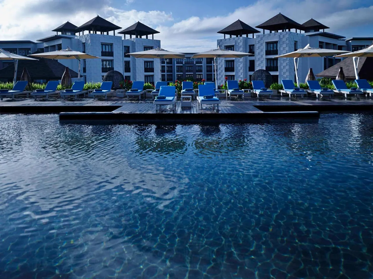A large blue swimming pool with rows of blue sun loungers and parasol umbrellas on the deck, with multi-storey hotel buildings with pavilion roofs behind.