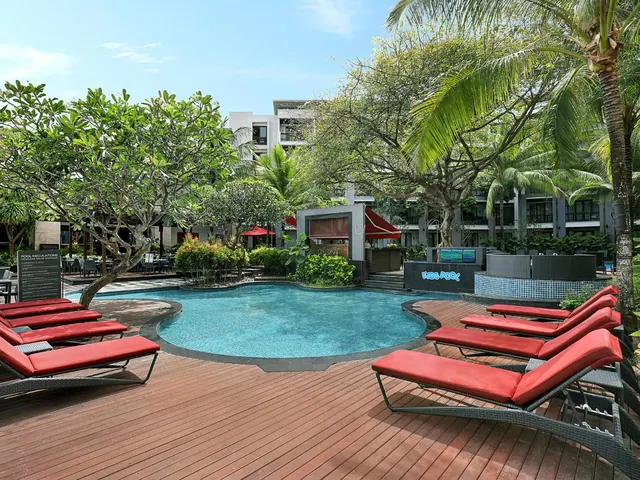 A kidney-shaped swimming pool with red-cushioned sun loungers on a timber deck, a pool bar and a Kids Pool sign beyond, surrounded by palm and frangipani trees.