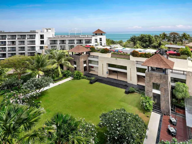 Aerial view of the hotel buildings and a lawn lined with palm trees, with the sea visible in the background.