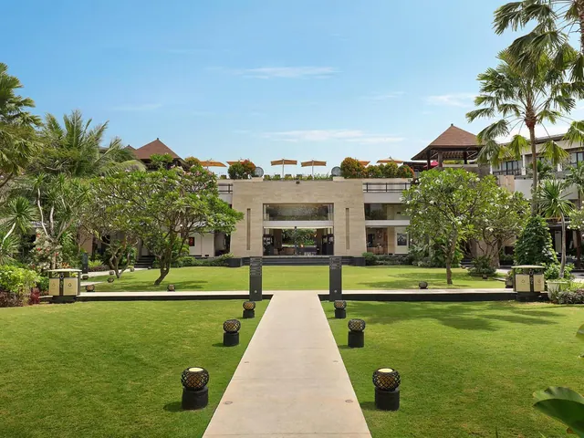 A manicured lawn with a concrete pathway leads toward a stone-faced hotel building, lined with lanterns and frangipani trees.