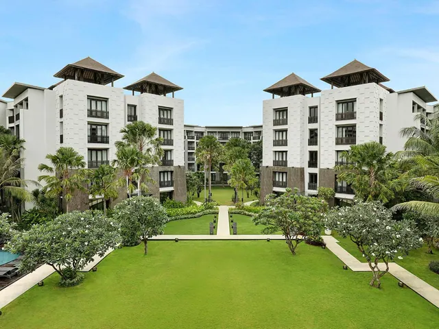 The exterior of a white multi-storey resort building with pagoda-style roofs stands behind a lawn lined with frangipani trees and palms.