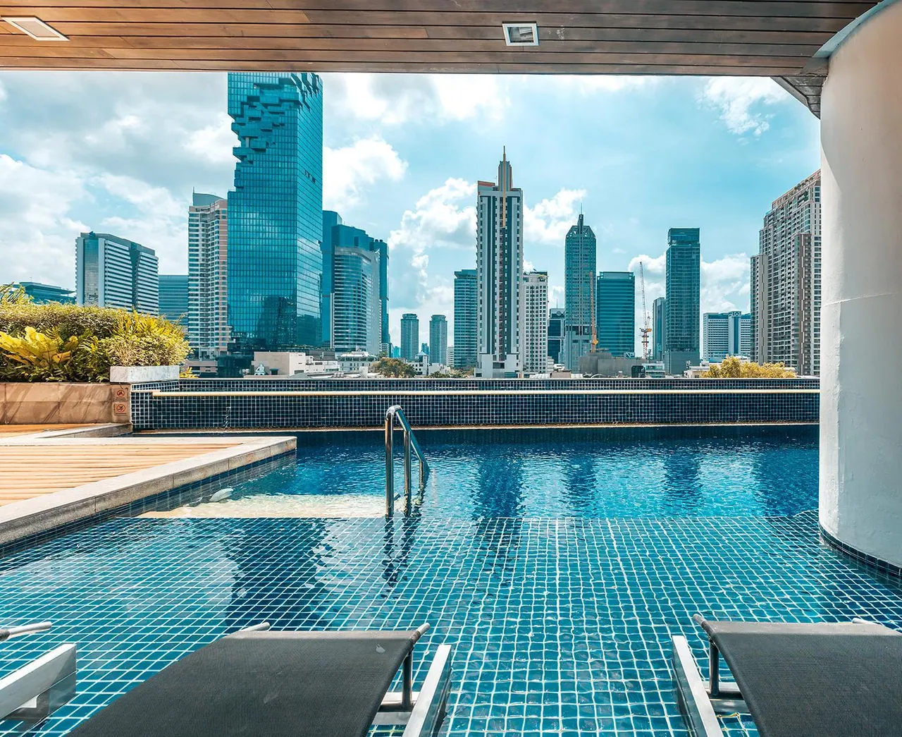 A rooftop swimming pool with blue mosaic tiles and sun loungers in the foreground, looking out over a city skyline of high-rise towers.