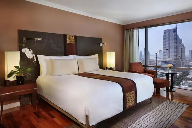 A bedroom with a made king bed, a red armchair and a round side table beside floor-to-ceiling windows overlooking city high-rises.