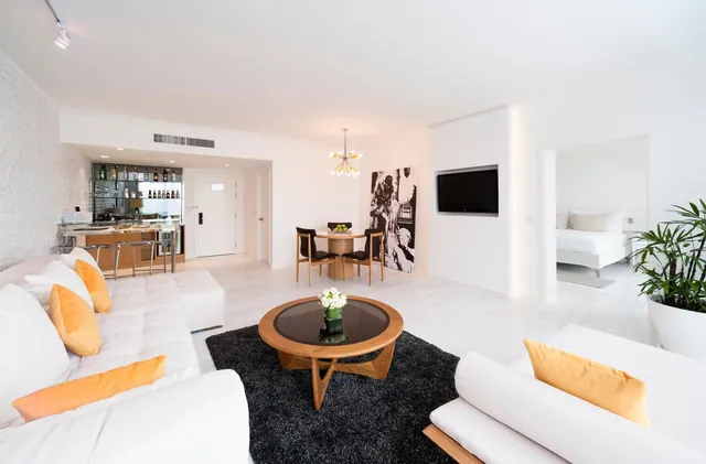 A white living area with sofas, a round wooden coffee table on a black rug, a dining table, a wall-mounted television and a bed visible through an open doorway.