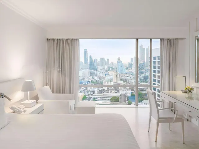 A white hotel bedroom with a large bed, an armchair and a desk, facing floor-to-ceiling windows that look out over a city skyline.