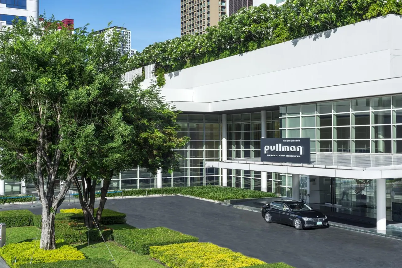 Exterior of a Pullman hotel entrance with a dark sedan parked under the canopy, a driveway, hedges and trees in front, and a vine-covered rooftop.