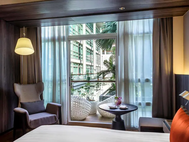 A hotel bedroom with a bed in the foreground, a wingback armchair beside floor-length curtains, and a glass door opening onto a balcony with wicker chairs and a view of palm trees in a courtyard.