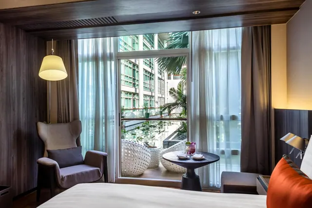 A hotel bedroom with a large bed and an armchair beside a tall window that looks out over a courtyard with palm trees and a multi-storey building.