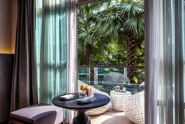 A round black table with fruit and napkins stands in a room beside an open glass door onto a balcony with white rattan chairs and palm trees.