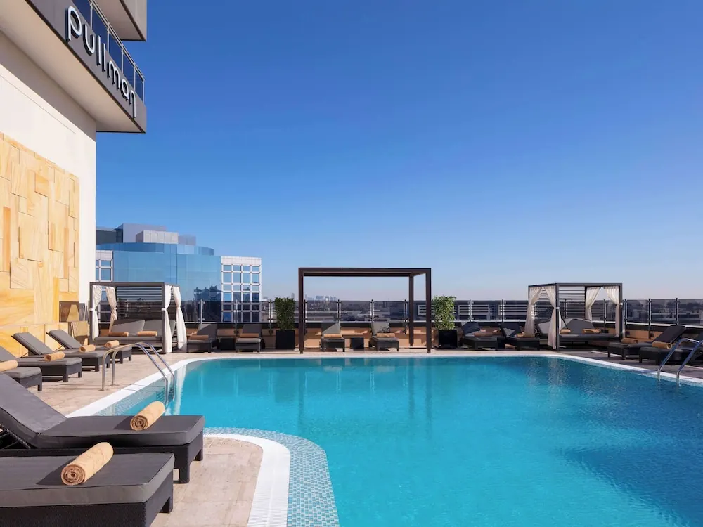 A rooftop swimming pool with sun loungers, curtained cabanas and a pergola, with a city skyline beyond the railing under a clear blue sky.