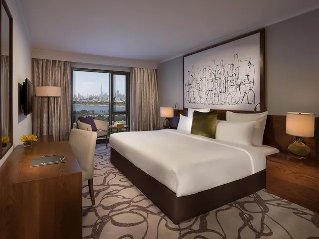 A king bed with white linen sits in a hotel bedroom beside a window overlooking a city skyline with the Burj Khalifa and a body of water.