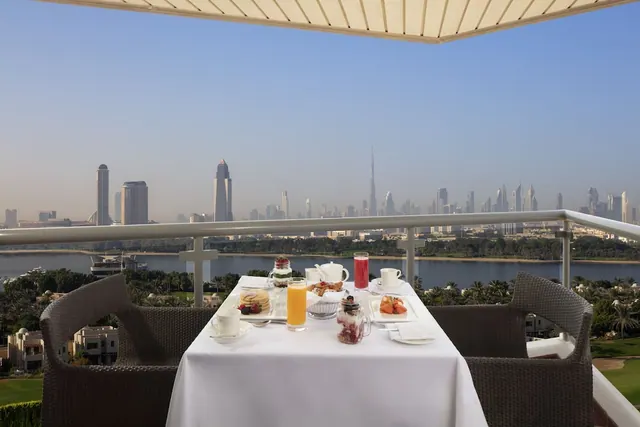 A breakfast table is set on a balcony overlooking a city skyline with the Burj Khalifa, a lake and green grounds below.