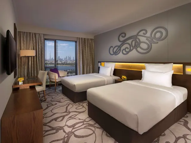 A twin bedroom with two made beds, a desk, a wall-mounted television and a window view of a city skyline including the Burj Khalifa across water.