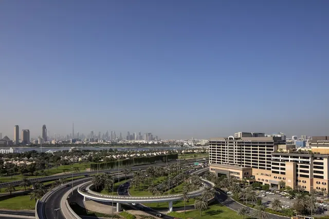 View from an upper floor across a highway interchange and palm-lined road toward a hotel building, a body of water, and a distant city skyline with the Burj Khalifa.