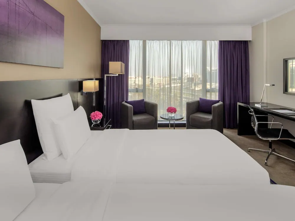 A hotel bedroom with a large white bed, two grey armchairs by tall windows looking out over city buildings, and a desk with a chair on the right.