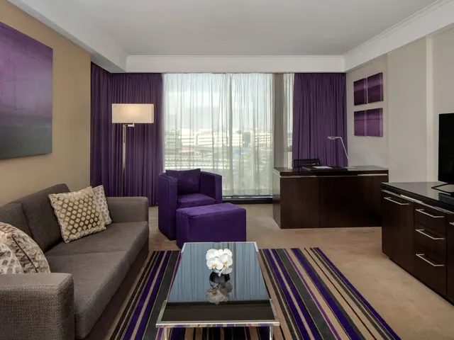 A hotel suite living area with a grey sofa, a purple armchair and ottoman, a coffee table with an orchid, a desk, a television, and a window onto low city buildings.