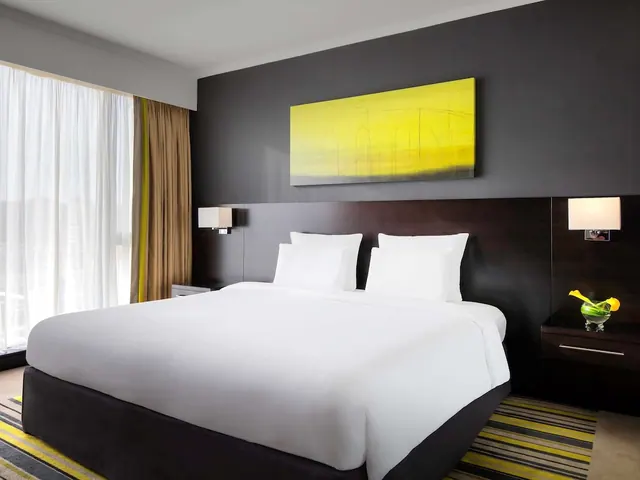 A bedroom with a large white bed, a dark wood headboard with wall lamps, a yellow wall painting, and a window with sheer curtains.