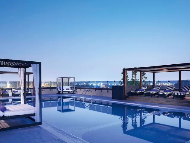 A rooftop swimming pool at dusk with white cabana beds, striped sun loungers and city lights visible beyond the glass railing.